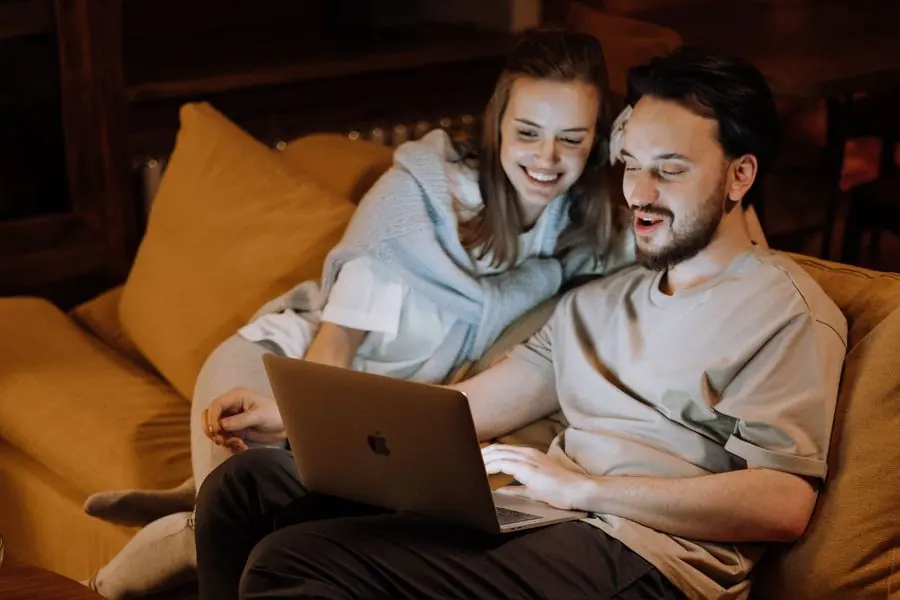 Un bărbat și o femeie stau pe canapea și se uită zâmbind la ecranul unui laptop.